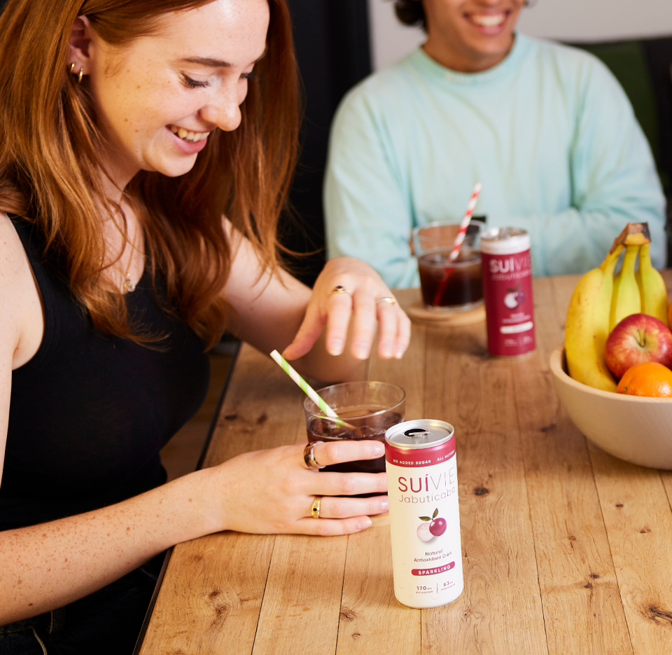 Woman drinking Suivie Jabuticaba from a glass with a straw, surrounded by fruits and cans on a wooden table.