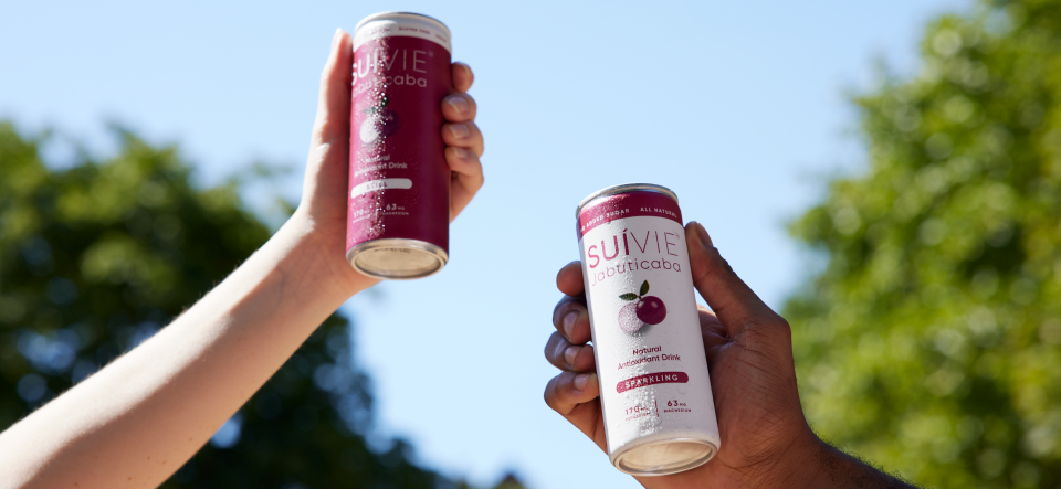 Two hands holding cans of Suivie Jabuticaba against a blurred outdoor background