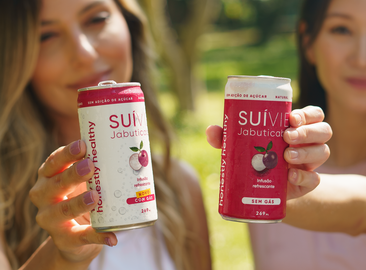 Two women enjoying Suivie Jabuticaba cans outdoors with greenery in the background